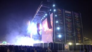 Veduta notturna di un grande palco al Sziget Festival. Faretti e schermi LED illuminano il palco, mentre una fitta folla si raduna davanti. Sul lato della struttura del palco sono appesi grandi banner con il logo di Sziget e i nomi di vari artisti.